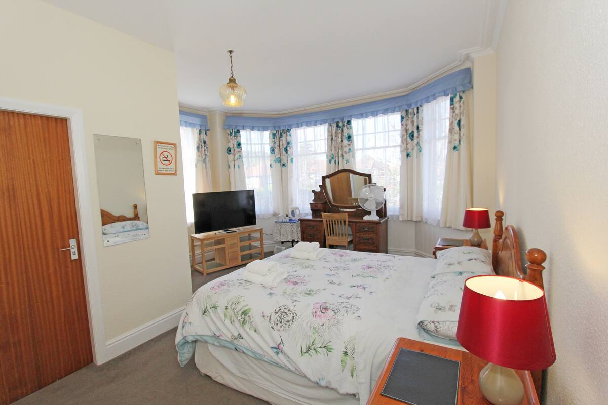 A bedroom with a bed, a desk, a chair, a television, a mirror, and a lamp.