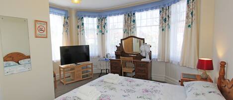 A bedroom with a bed, a desk, a television, a mirror, and a nightstand with a lamp.