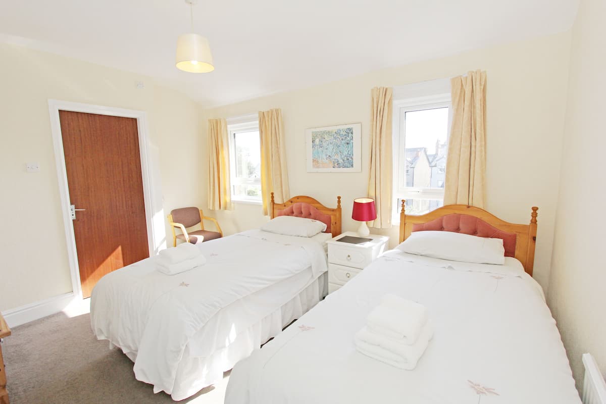 A hotel room with two beds, a wooden headboard, a nightstand with a lamp, a chair, and a door.