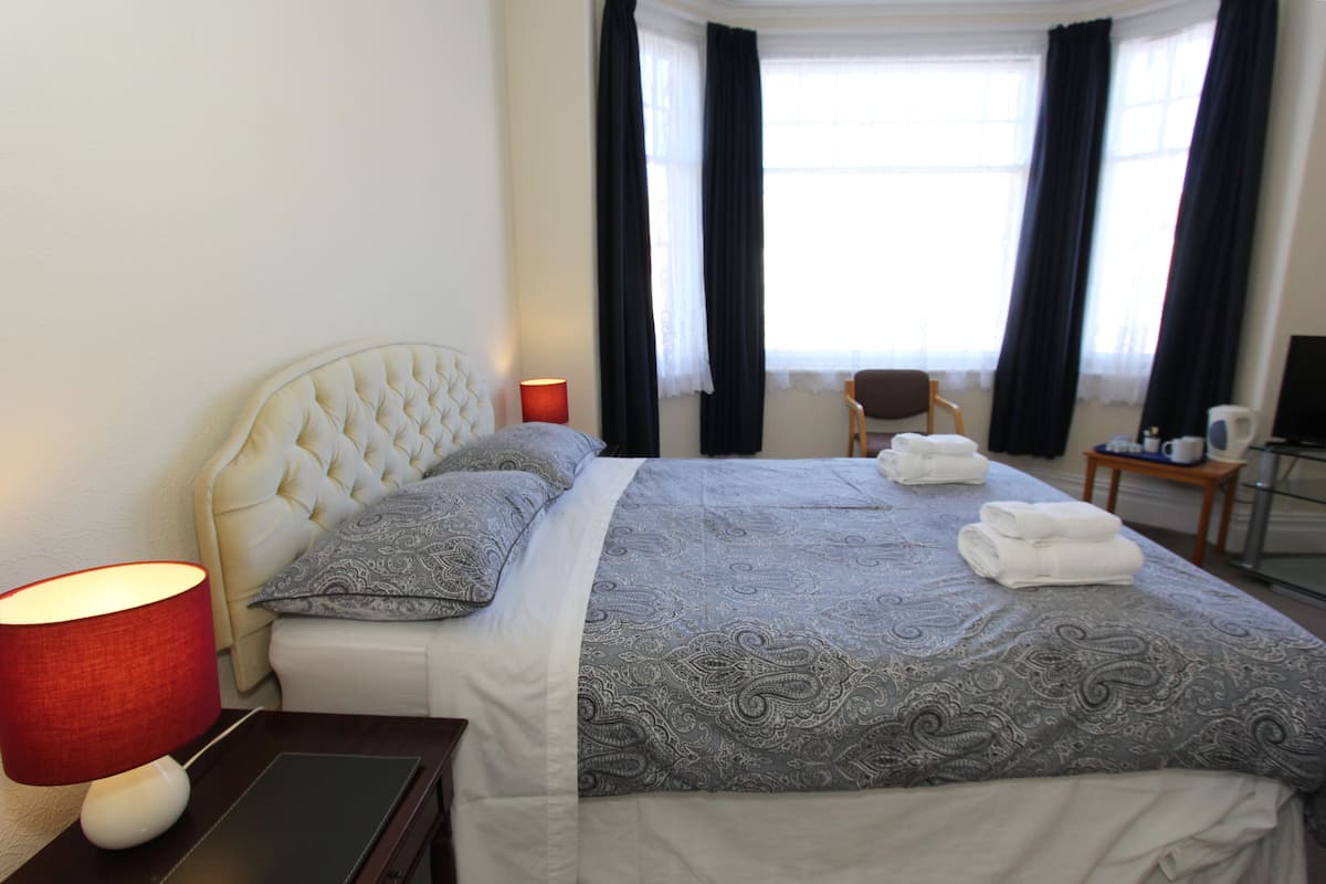 A hotel room with a neatly made bed, a bedside lamp, a chair, and a desk with a computer.