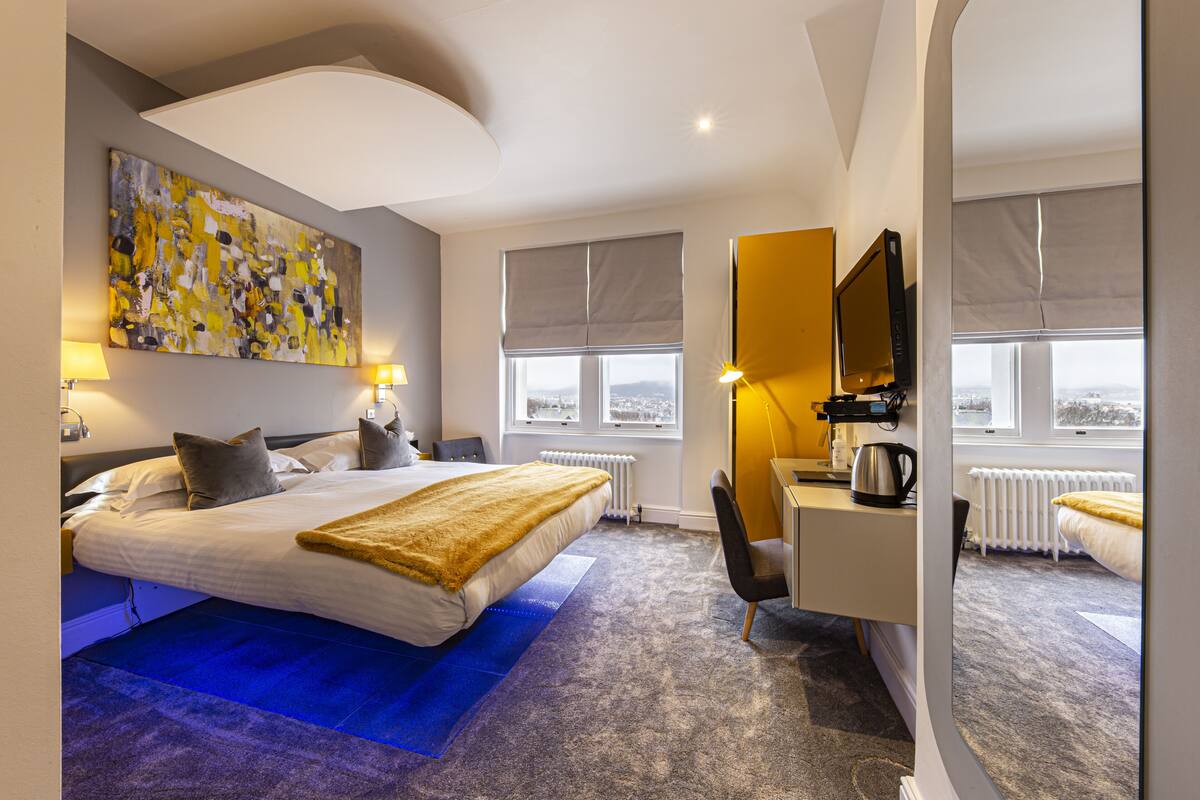 A modern hotel room with a large bed, a desk, a chair, and a large window with a view.
