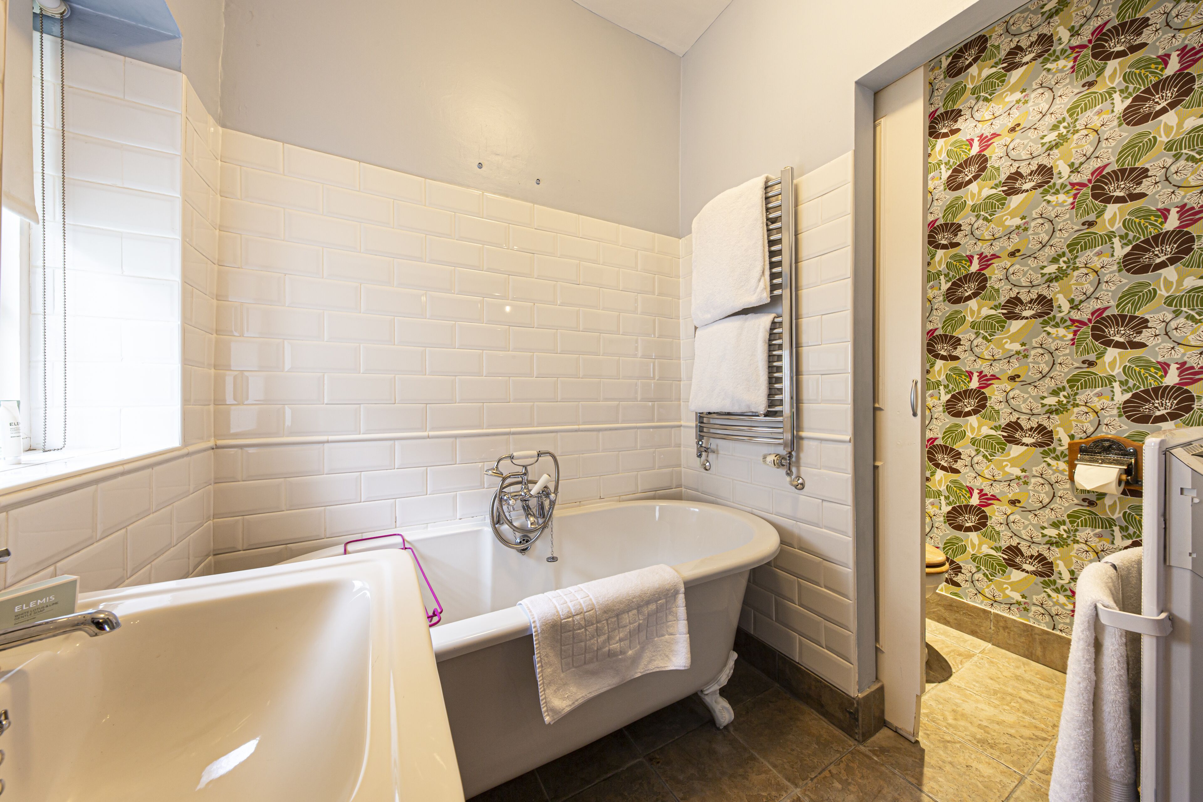 Um banheiro com parede revestida de azulejos brancos, uma banheira livre, um porta-toalhas e papel de parede com padrão floral.