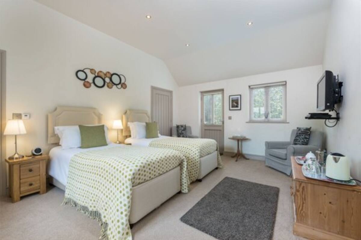 A bedroom with two beds, a bedside table with a lamp, a small table, a grey armchair, and a television mounted on the wall.