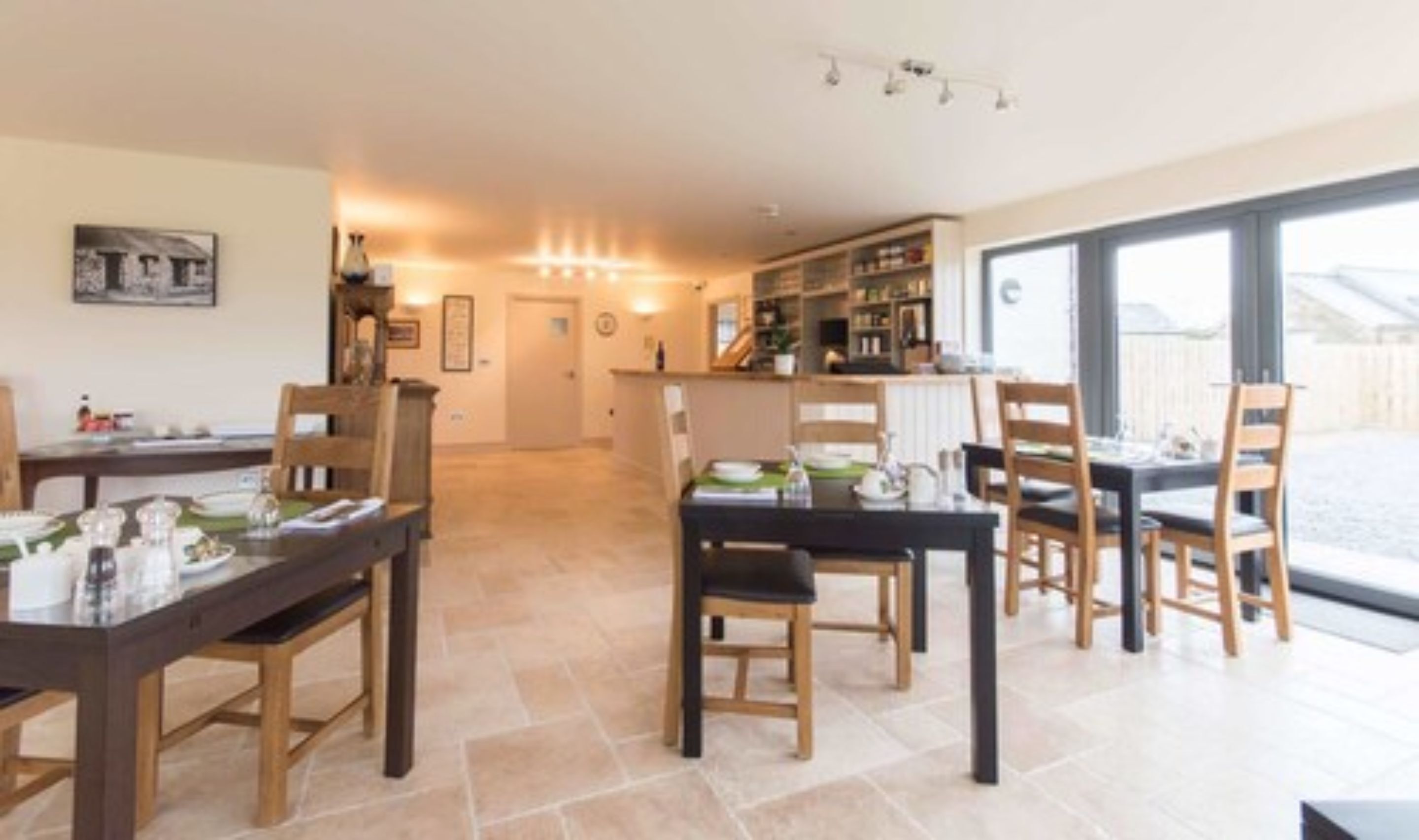 Un comedor amplio con mesas y sillas de madera, una cocina al fondo y grandes ventanales que dejan entrar luz natural.