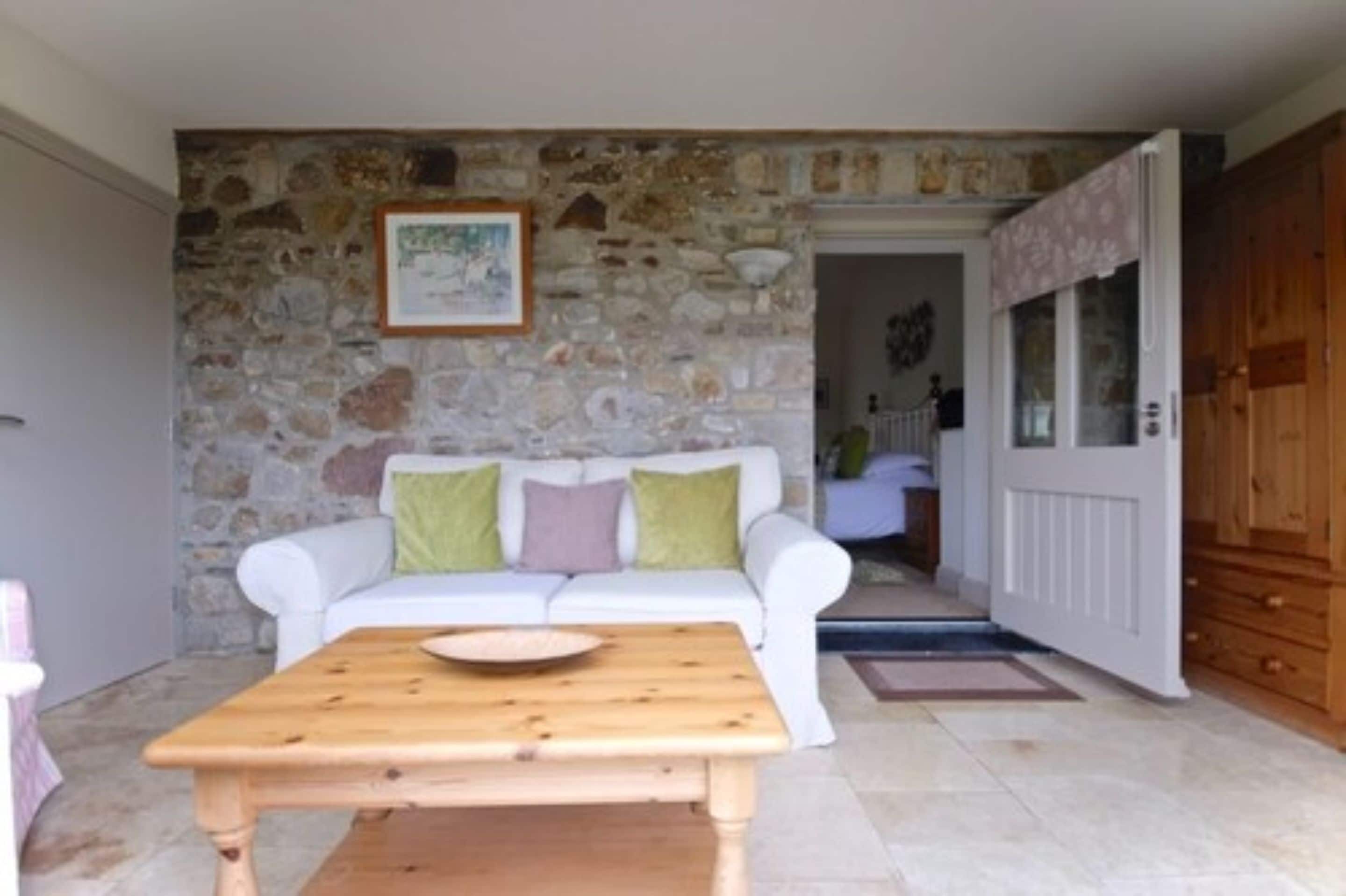 Una habitación con pared de piedra, sofá blanco, mesa de madera y una puerta que da a otra habitación.