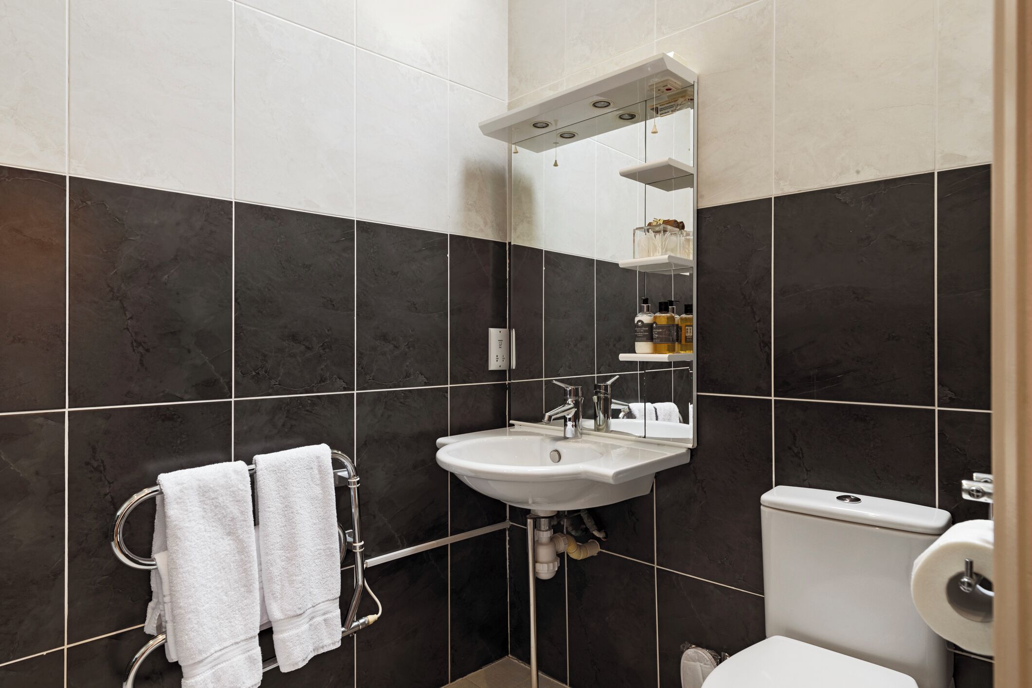 Un baño moderno con baldosas oscuras, un lavamanos blanco, un inodoro y un perchero con dos toallas.