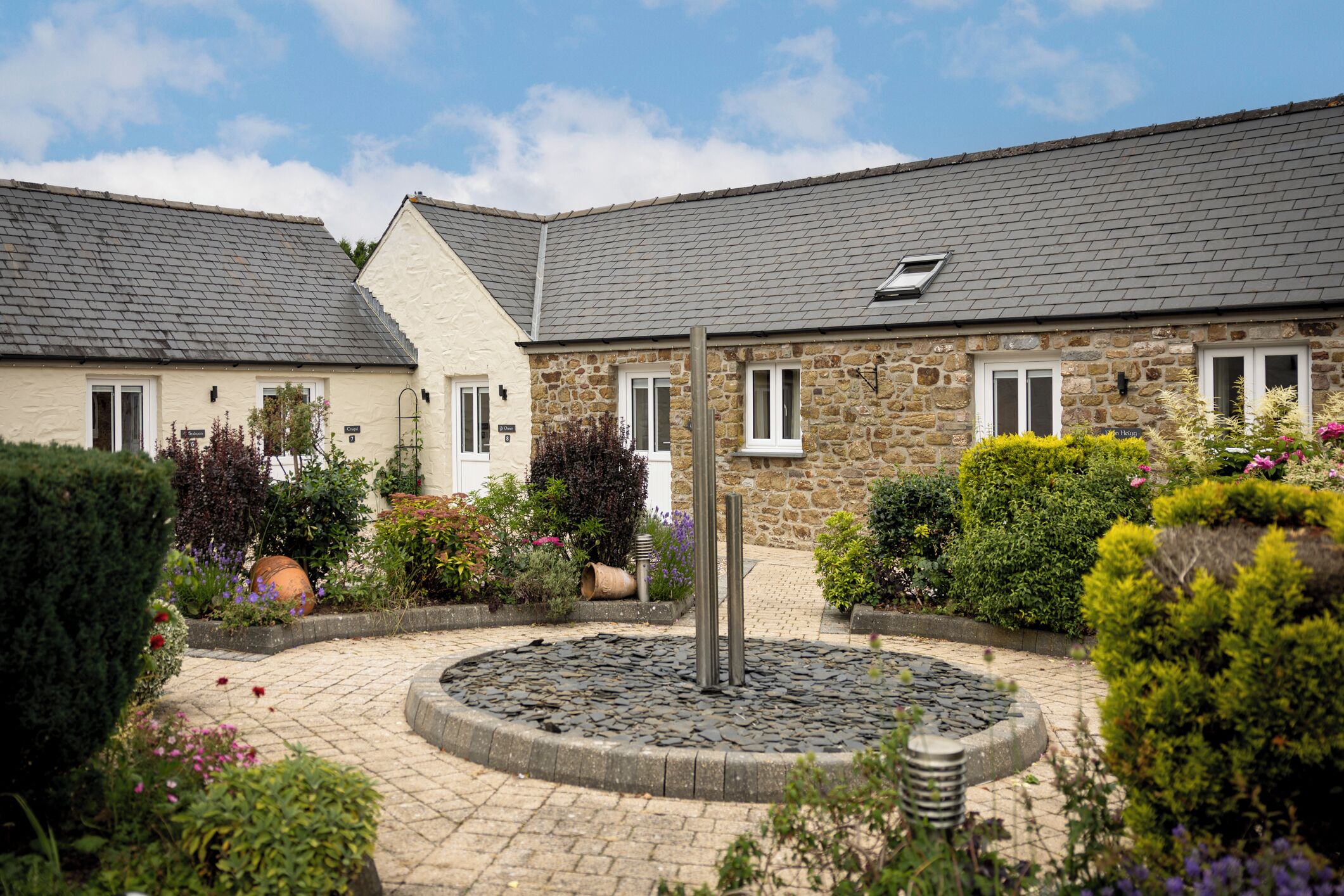 A courtyard with a central water feature, surrounded by stone buildings with windows and a variety of plants.