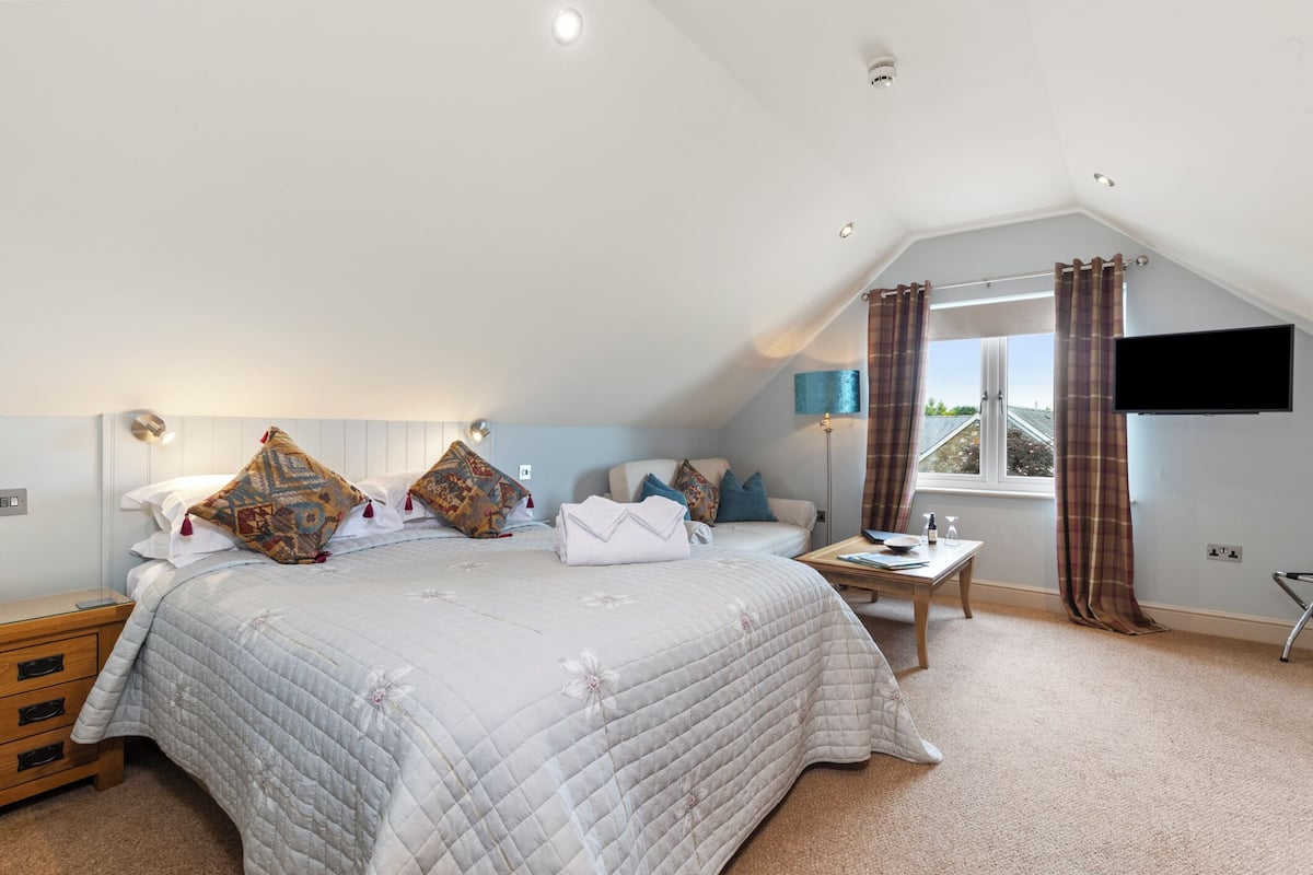 A bedroom with a large bed, a bedside table, a flat-screen TV mounted on the wall, and a window with curtains.