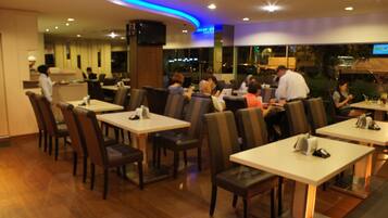Um restaurante com várias mesas e cadeiras, um balcão e uma tela de televisão.