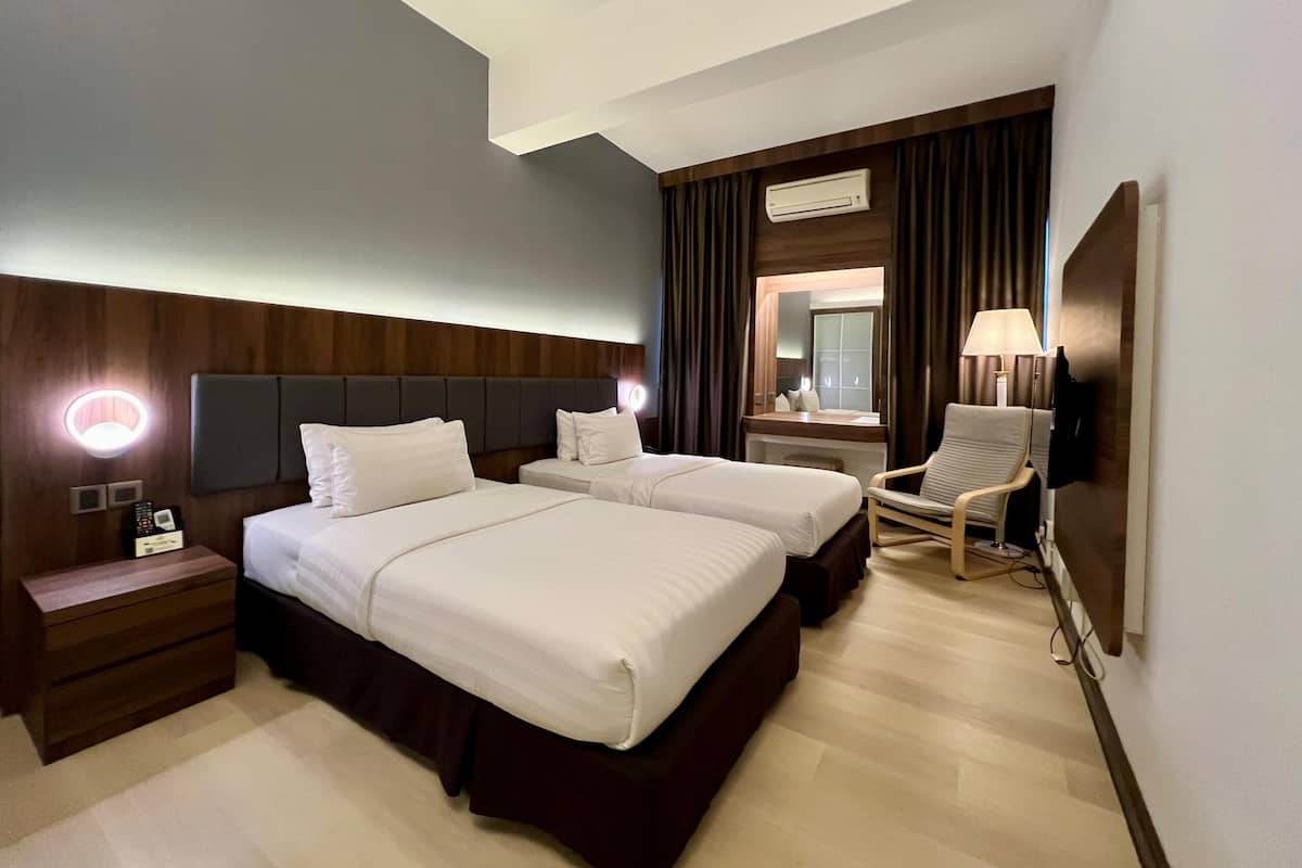 A hotel room with two beds, a desk, a chair, a bedside table, a lamp, and an air conditioning unit.