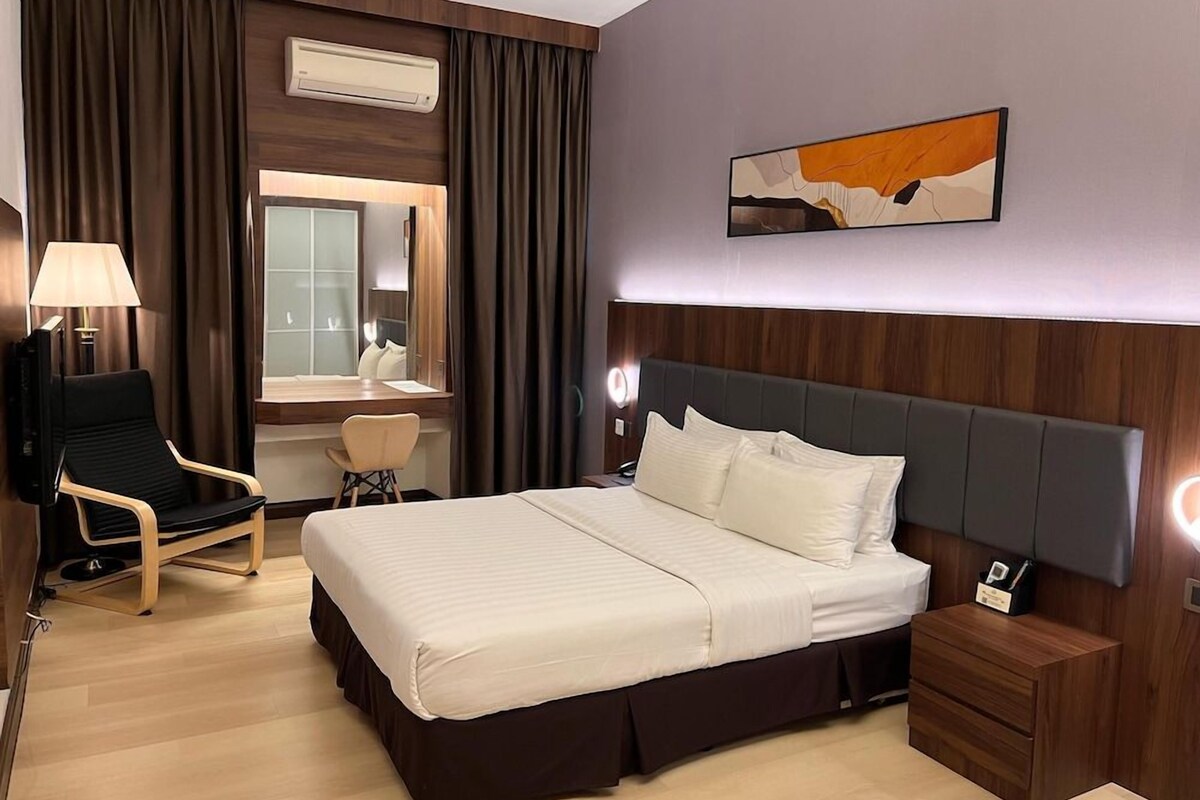 A hotel room with a bed, bedside table, chair, desk, and an air conditioning unit.