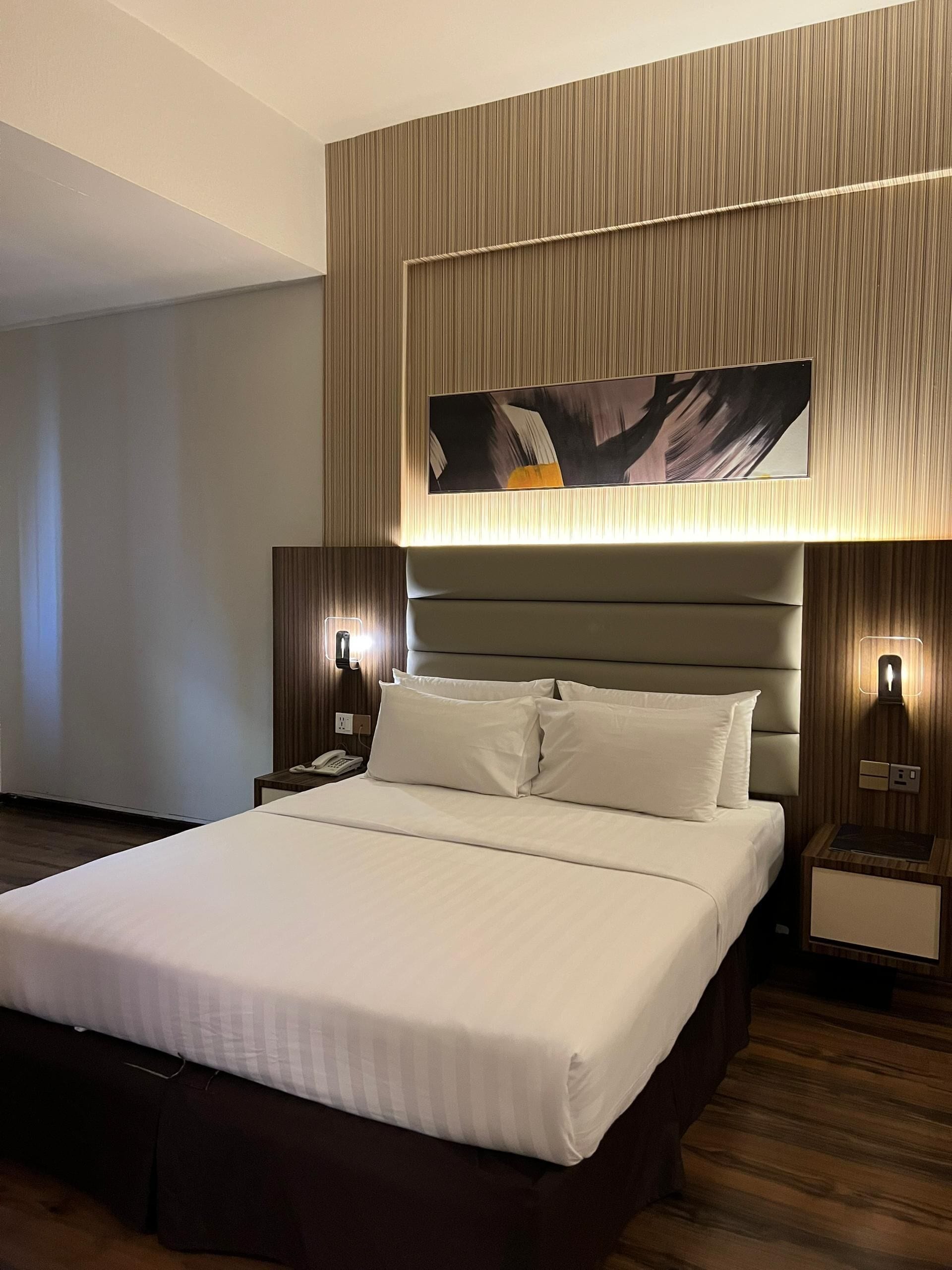 A modern hotel room with a large bed, a headboard with vertical lines, a painting above the bed, and two bedside lamps.