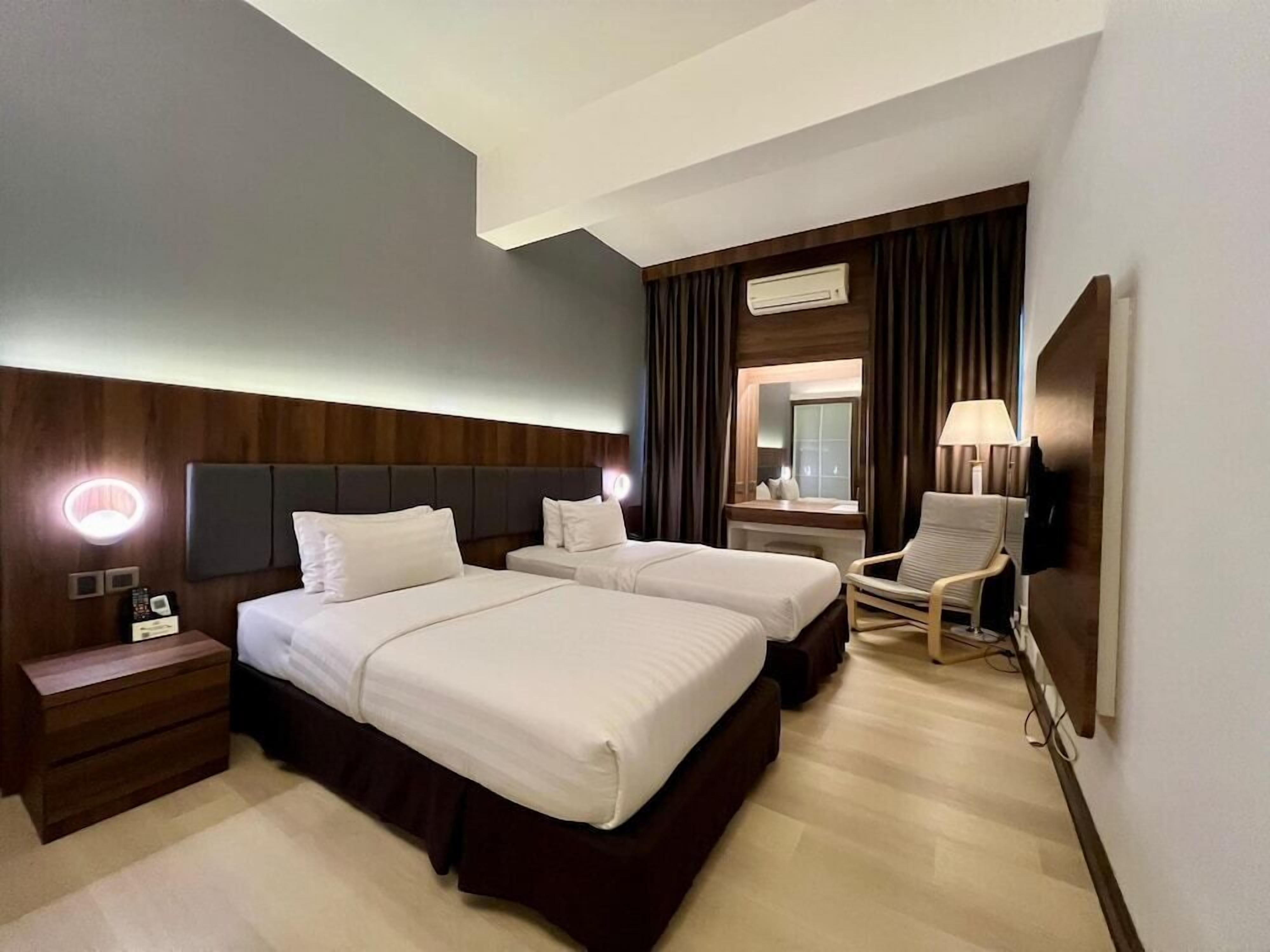 A hotel room with two beds, a nightstand, a chair, a desk with a lamp, and an air conditioning unit.