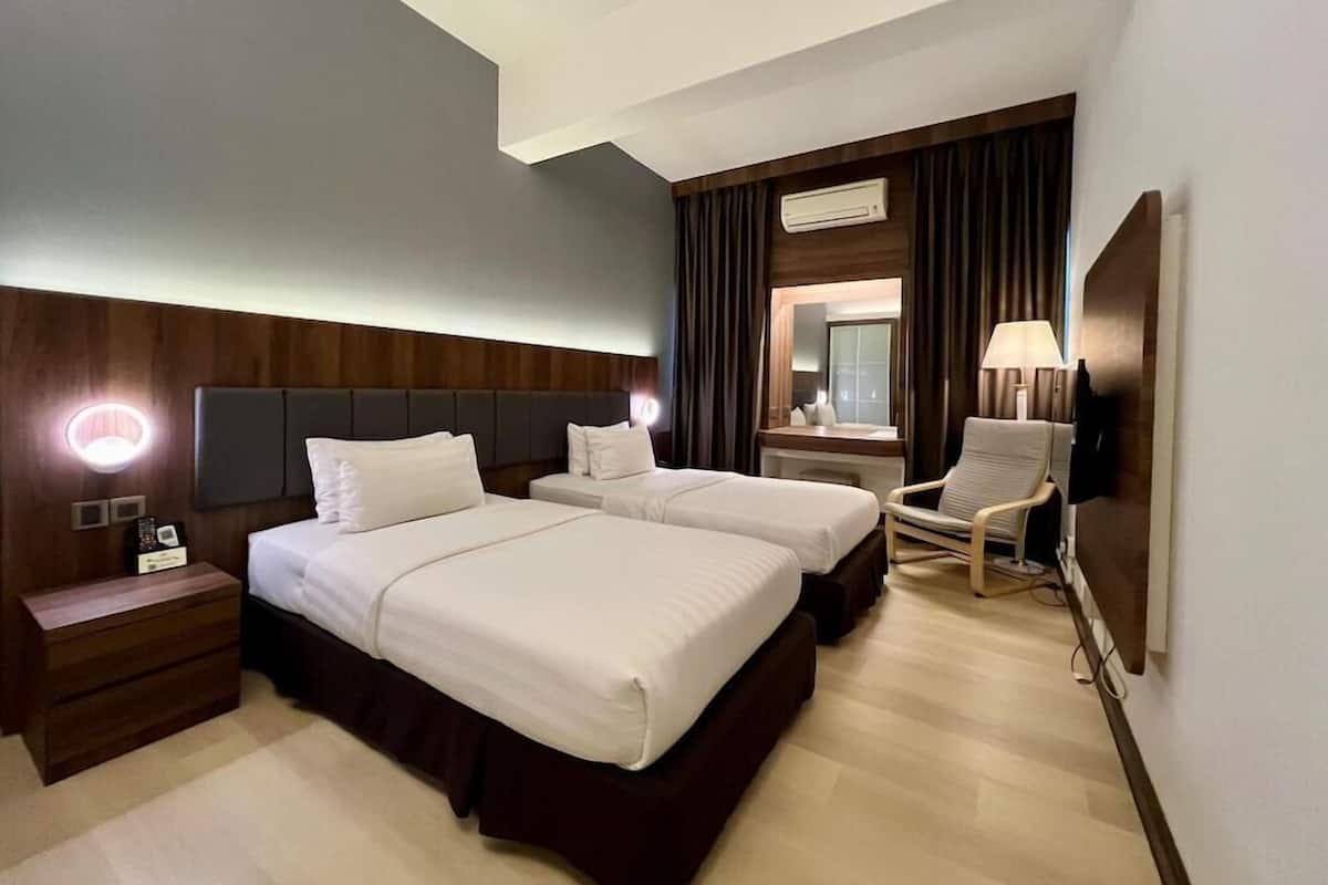 A hotel room with two beds, a nightstand, a chair, a desk with a lamp, and an air conditioning unit.