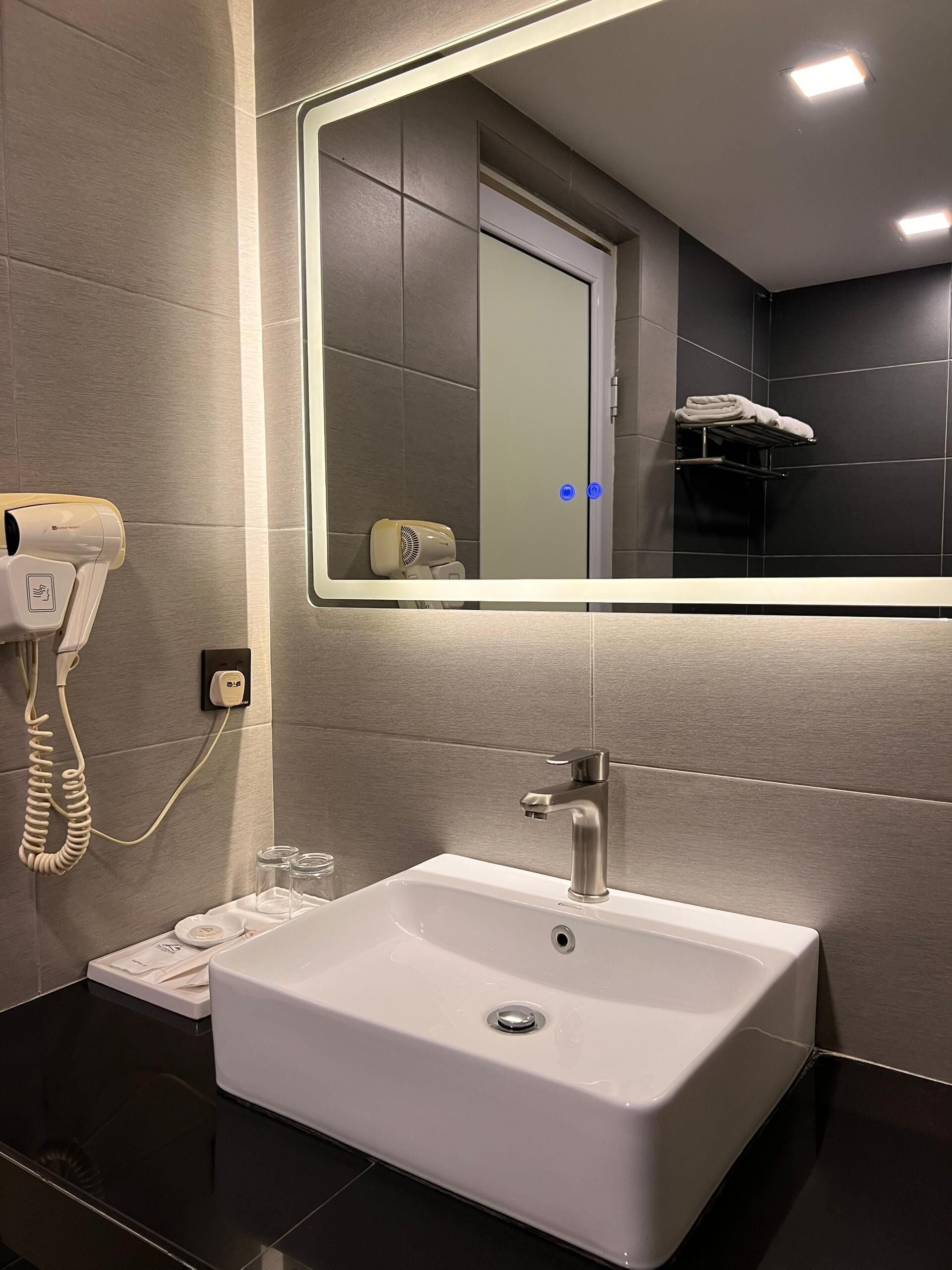 A modern bathroom with a large mirror, a white sink, a hairdryer, and a towel rack.