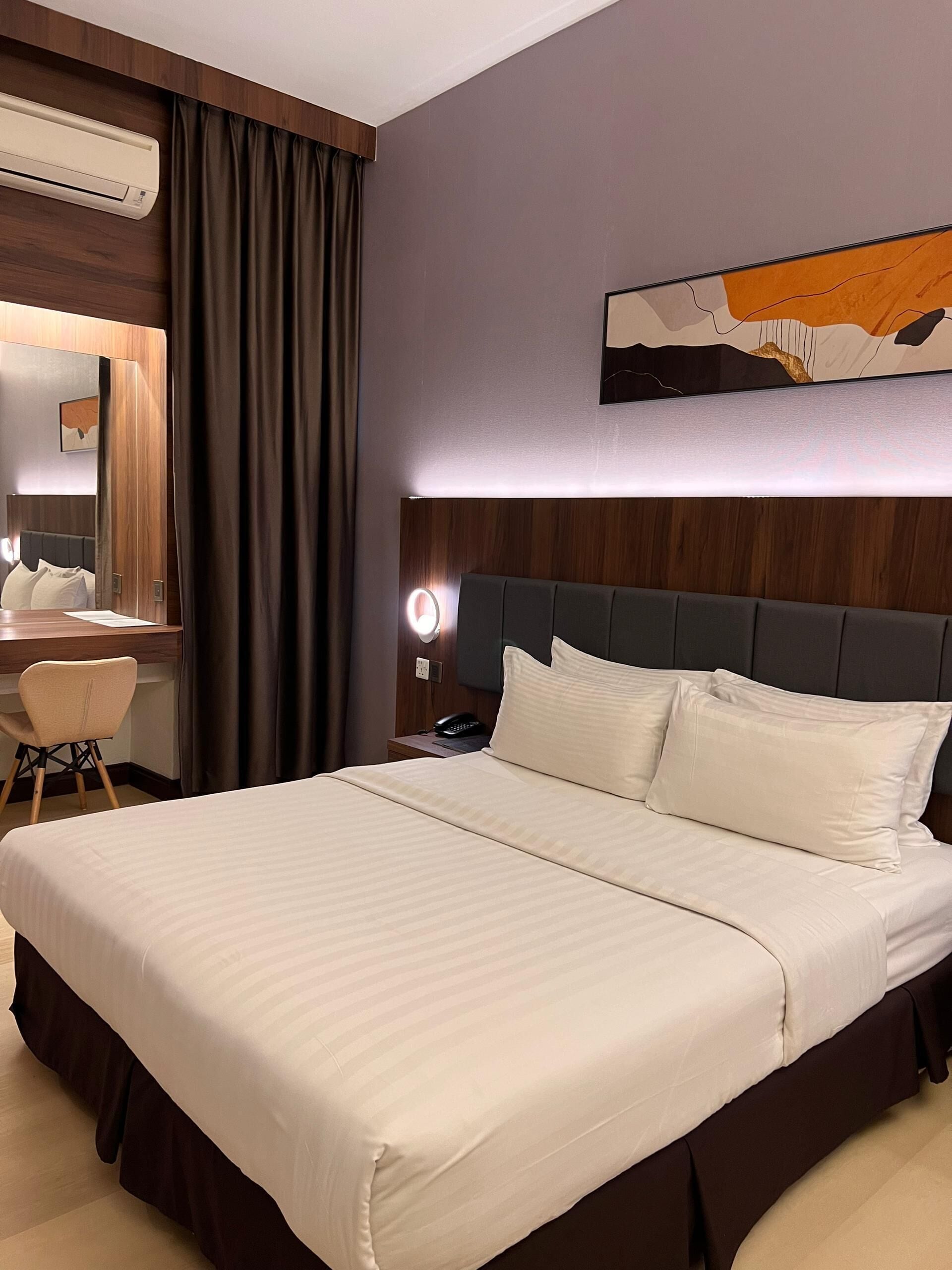 A hotel room with a large bed, a desk with a chair, a mirror, and a wall-mounted air conditioner.