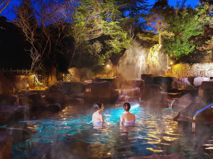 A hot spring area with two people in the water, surrounded by rocks and trees.