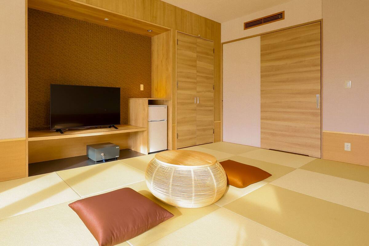 A modern hotel room with a television, a small refrigerator, and a futon bed with cushions.