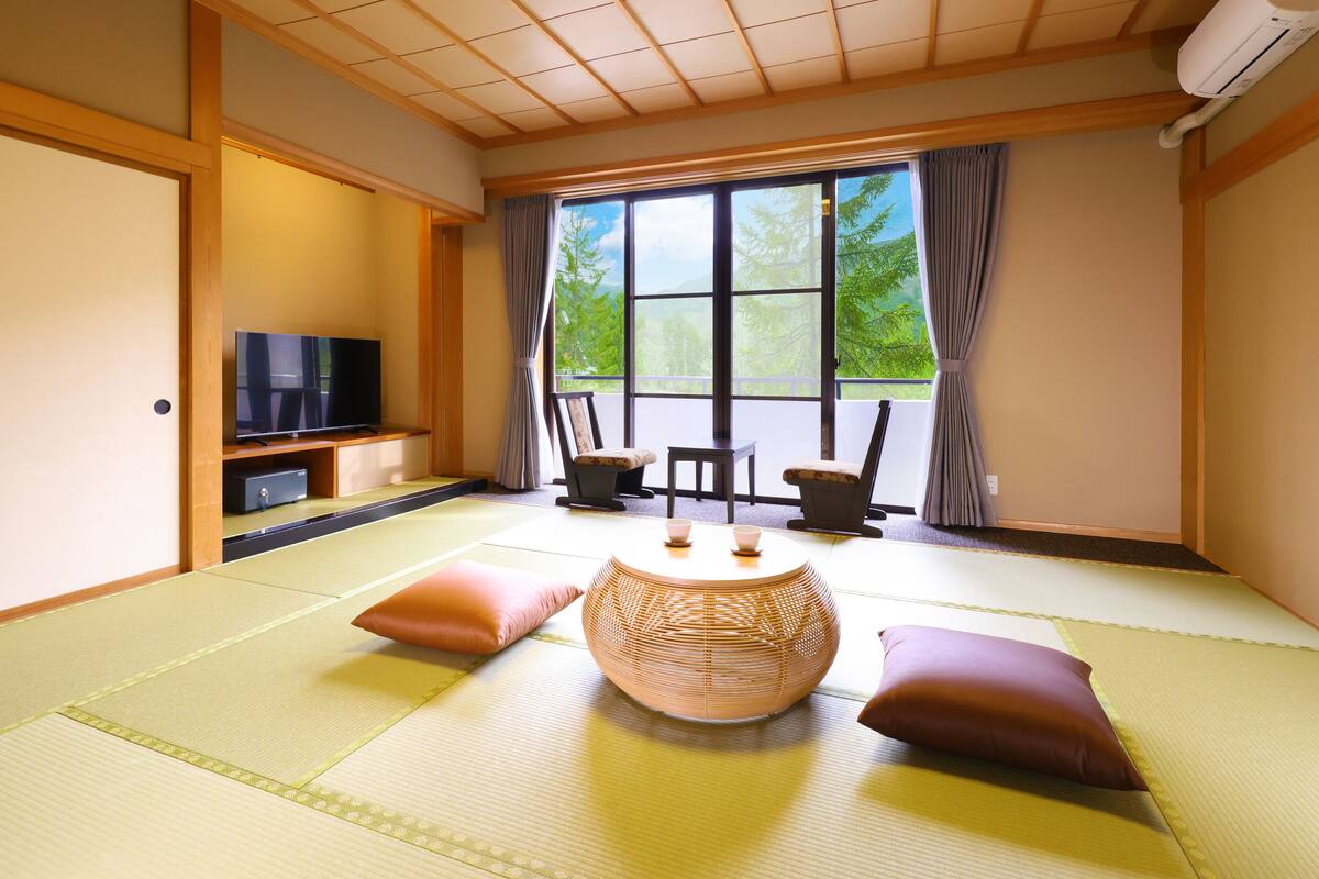 A traditional Japanese-style room with tatami flooring, a low table with cushions, a television, and a view of greenery through large windows.