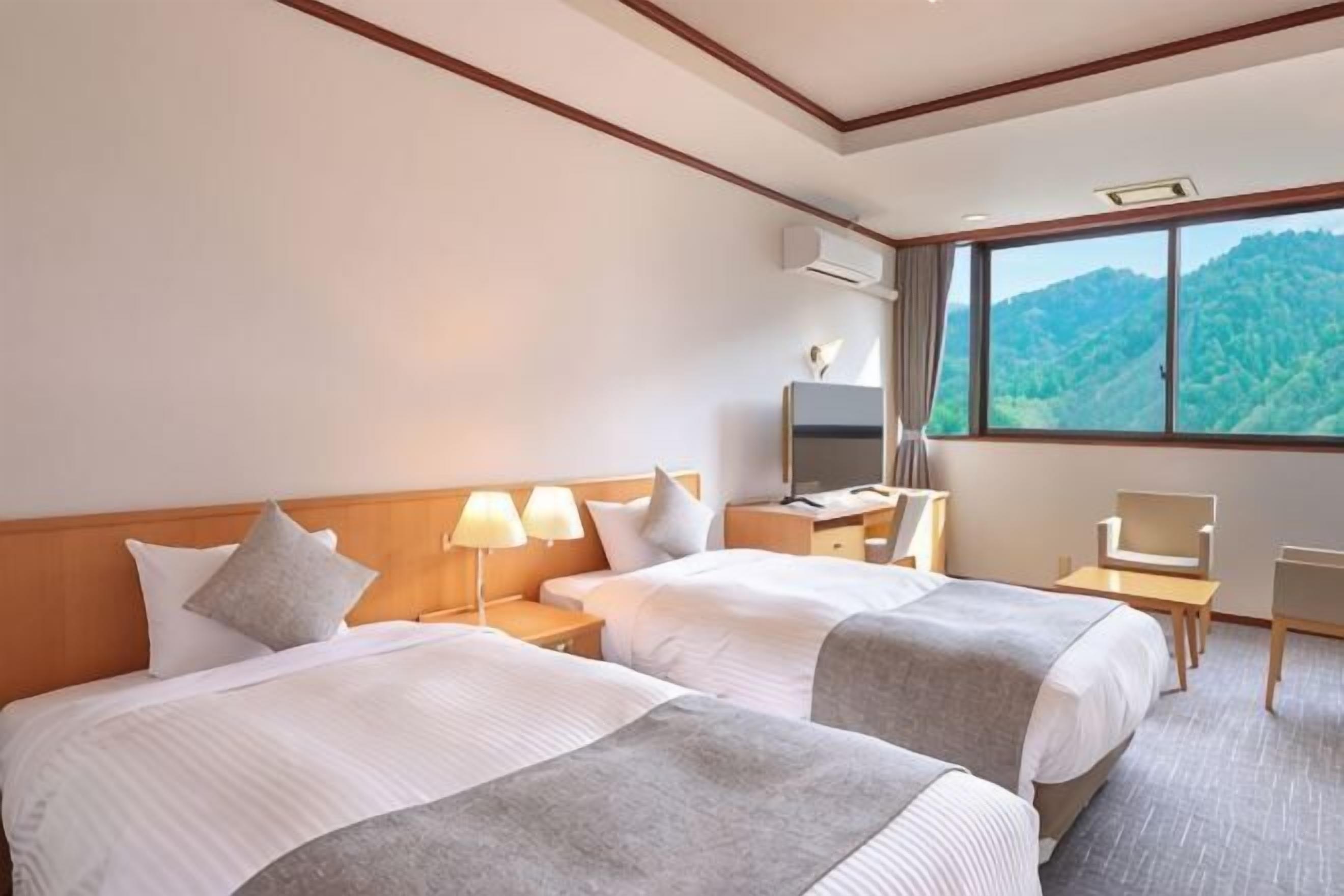 A hotel room with two beds, a desk with a chair, a TV, and a large window with a mountain view.