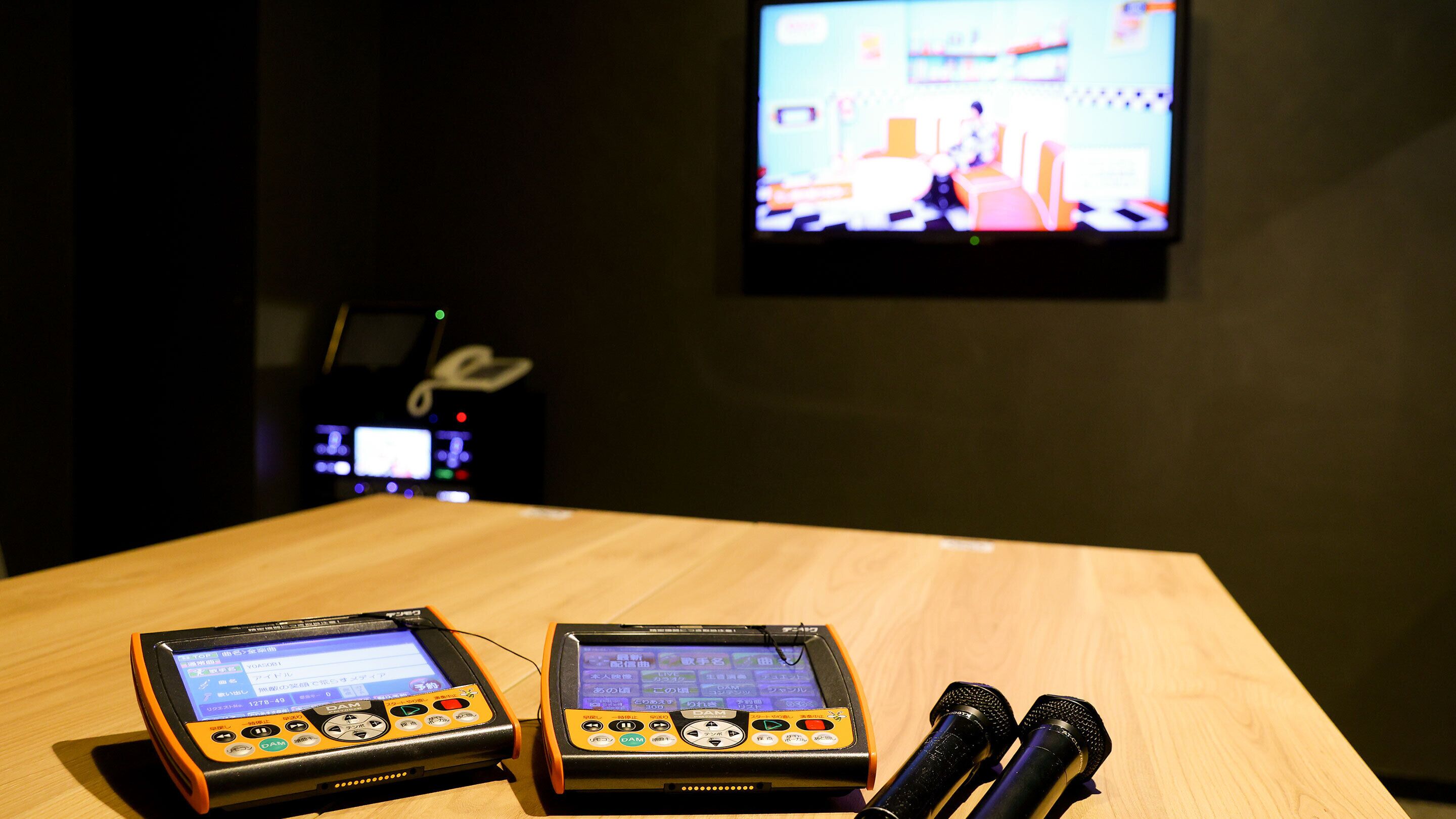 Two electronic devices on a wooden table, a pair of microphones, and a television in the background.