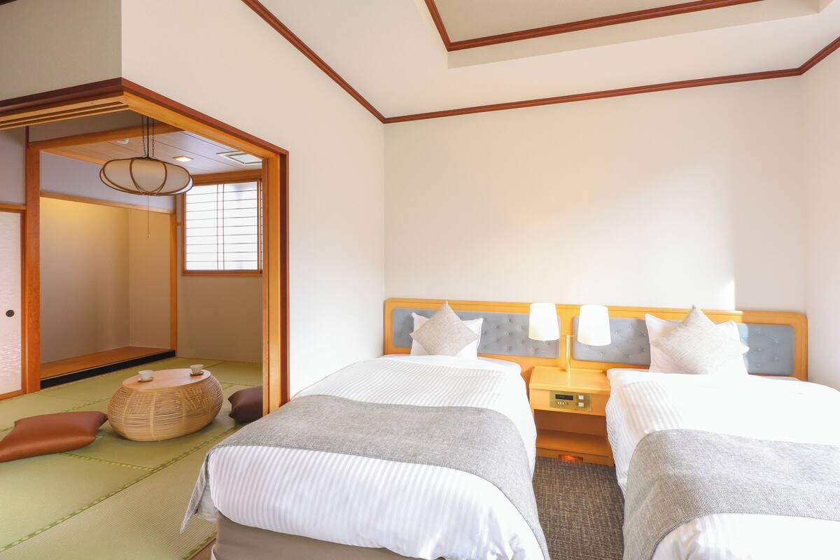 A hotel room with two beds, a traditional Japanese tatami mat floor, a wooden headboard, bedside tables with lamps, and a sliding door leading to a traditional Japanese room.