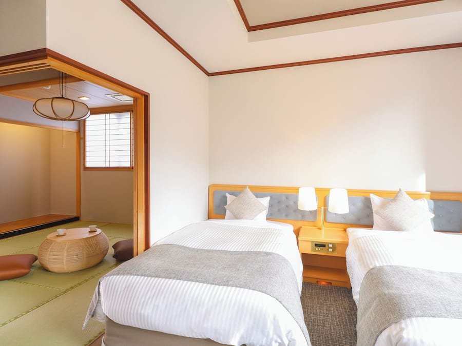 A hotel room with two beds, a traditional Japanese tatami mat floor, a wooden headboard, bedside tables with lamps, and a sliding door leading to a traditional Japanese room.