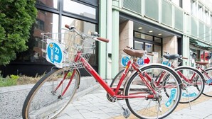 A row of red bicycles parked outside a building with a sign indicating a bike rental service. - Iidaya (Matsumoto)
