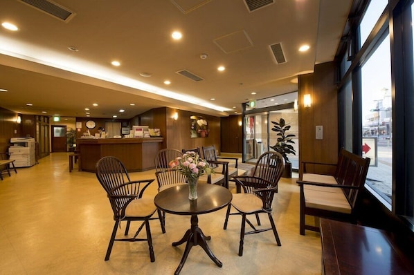 En hotellobby med en receptionsdisk, en sittgrupp med runda bord och stolar samt stora fönster med utsikt över gatan.