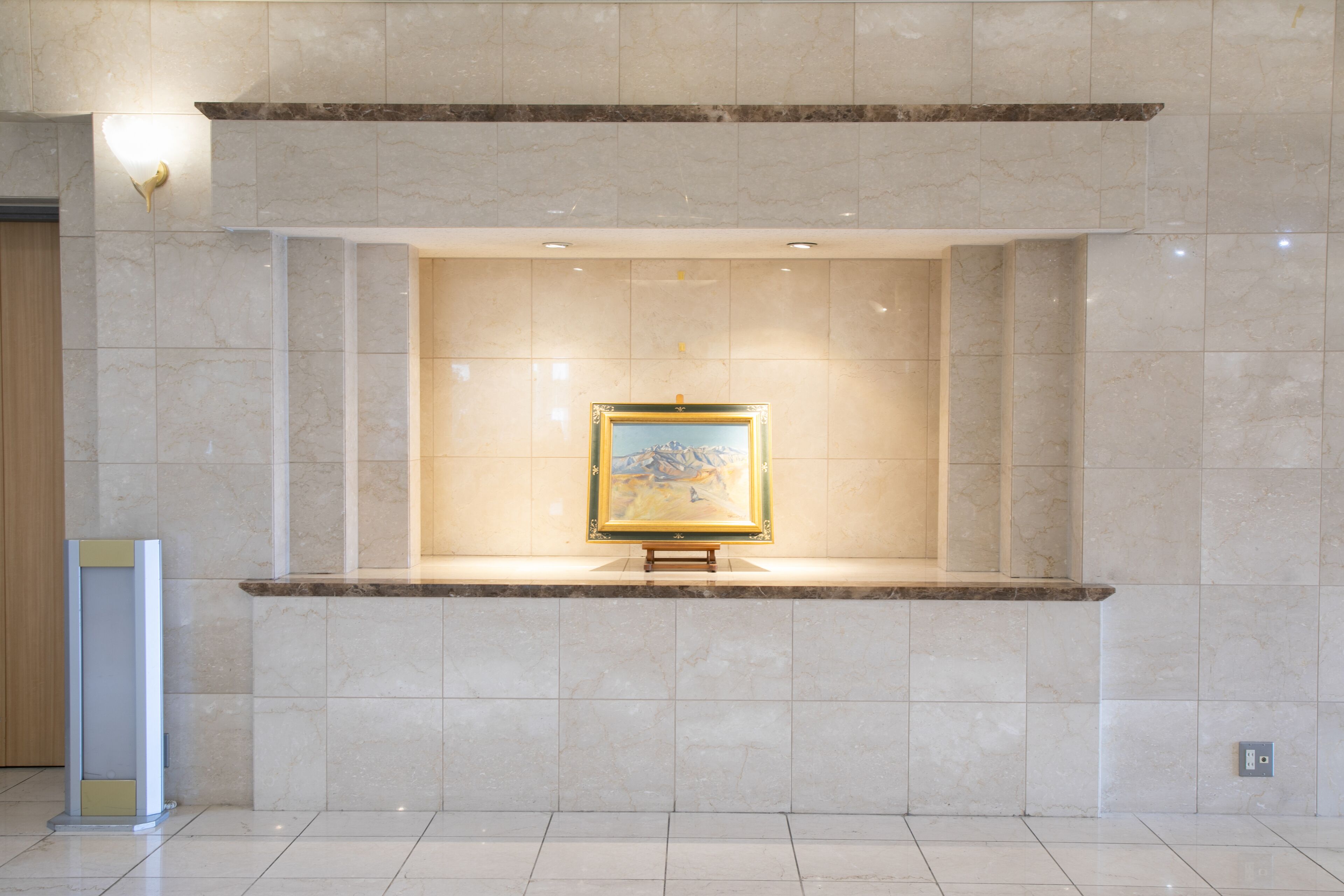 A lobby with a marble floor, a framed painting on a pedestal, and a light fixture above.
