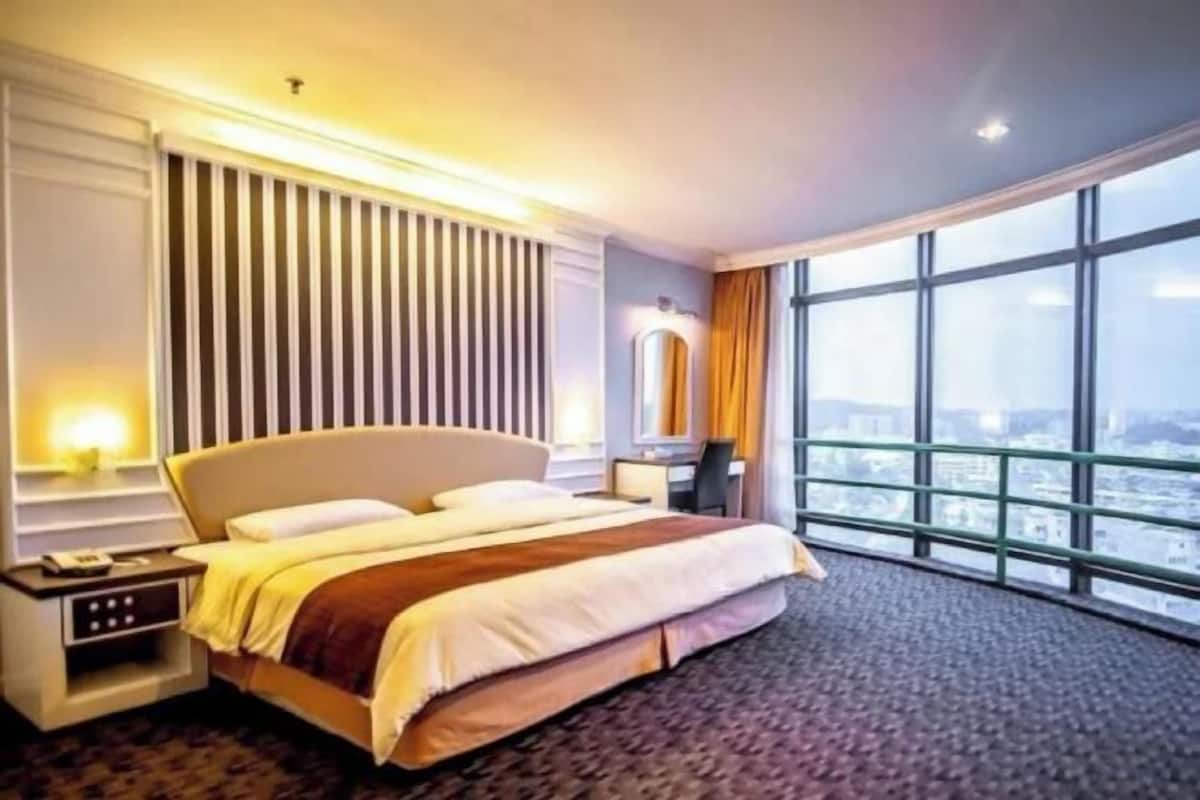 A hotel room with a large bed, a desk, a chair, a nightstand, and a view of the city through large windows.
