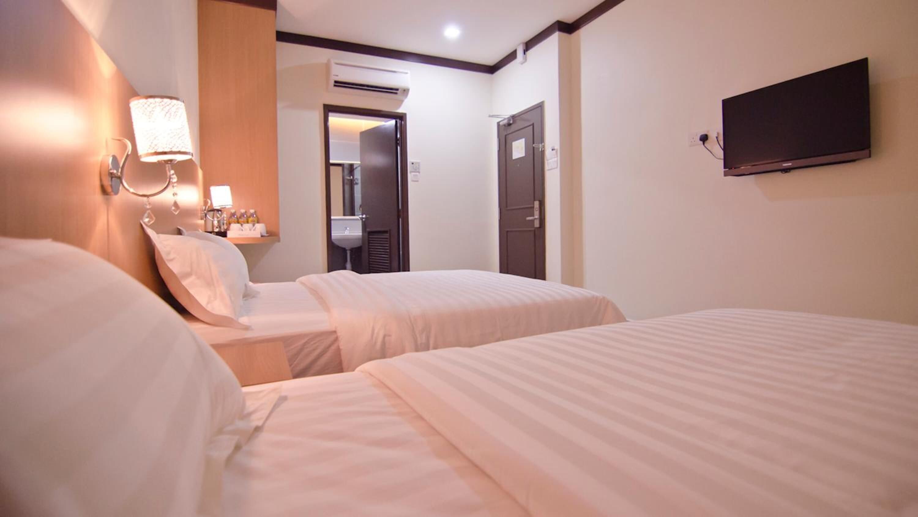 A hotel room with two beds, a wall-mounted TV, and a bathroom visible through an open door.
