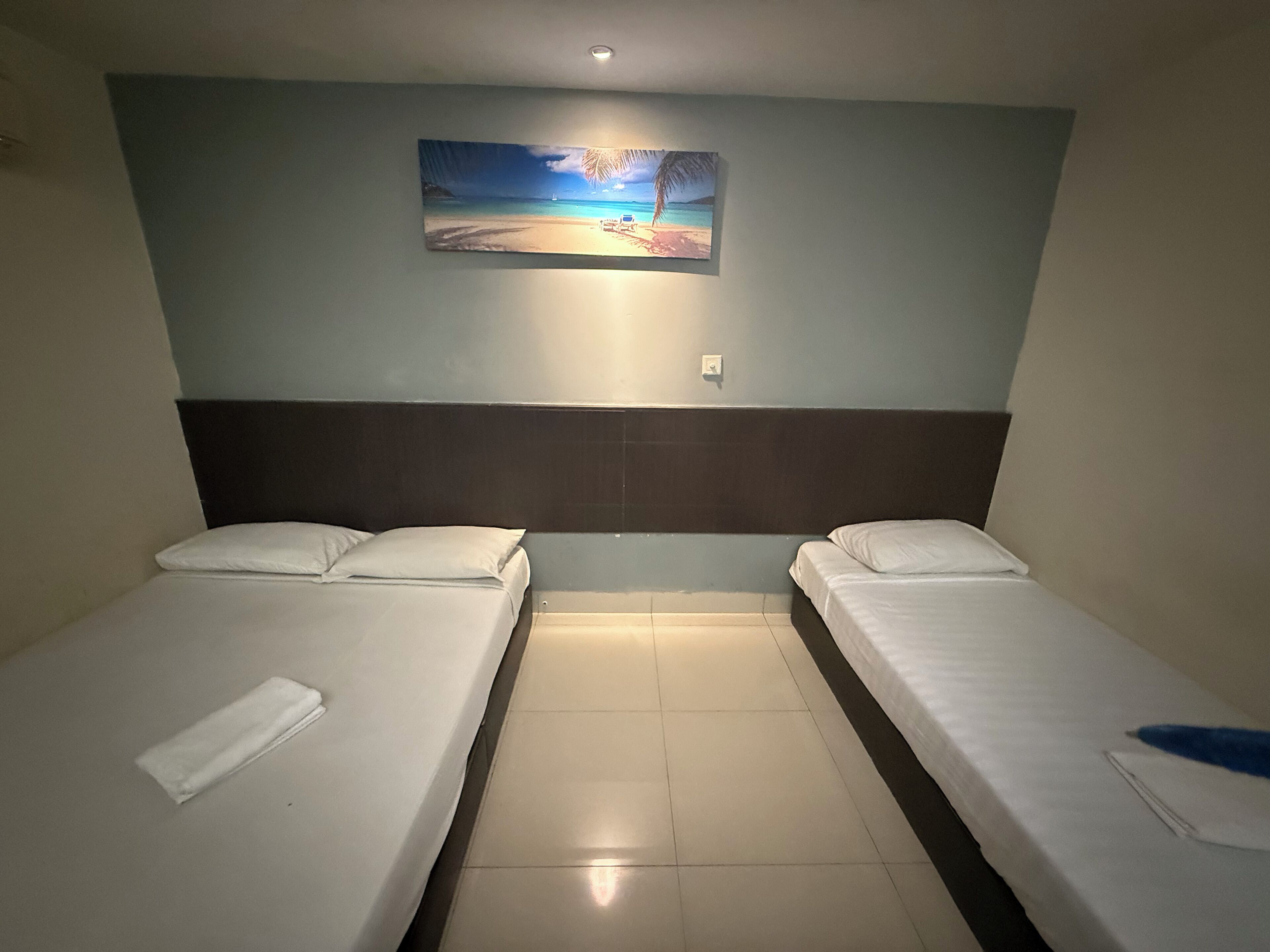 Habitación de hotel con dos camas individuales, una mesita de noche, un cuadro de una escena de playa y suelo de baldosas.