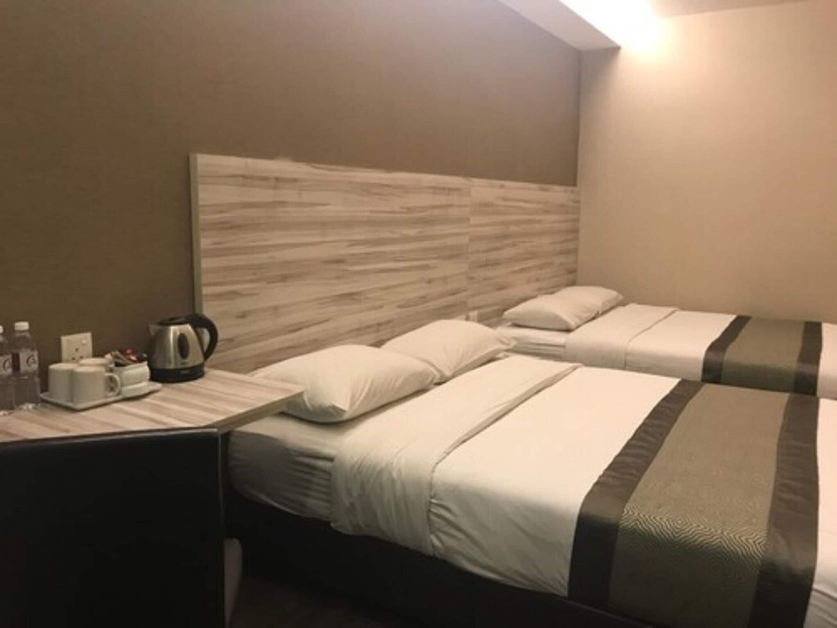 A hotel room with two single beds, a nightstand with a kettle and cups, and a wall-mounted headboard.