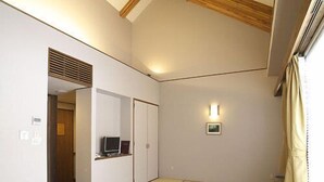 Une pièce avec des tatamis, une table en bois, des chaises, un téléviseur posé sur une étagère et une fenêtre avec des rideaux.