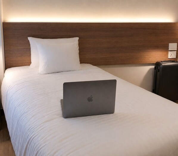 Cama bem arrumada com roupa de cama branca, um laptop sobre a cama e uma mala no chão.