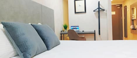 A hotel room with a bed, a desk with a laptop, a chair, a framed picture on the wall, and a coat hanger.