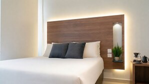 Una habitación de hotel moderna con una cama grande, cabecera de madera, mesita de noche con cafetera y una planta en maceta.
