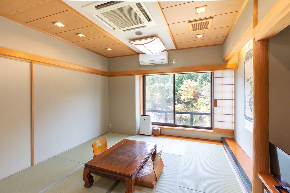 Una habitación con suelo de tatami, una mesa de madera, un televisor y un ventanal con vistas a árboles.