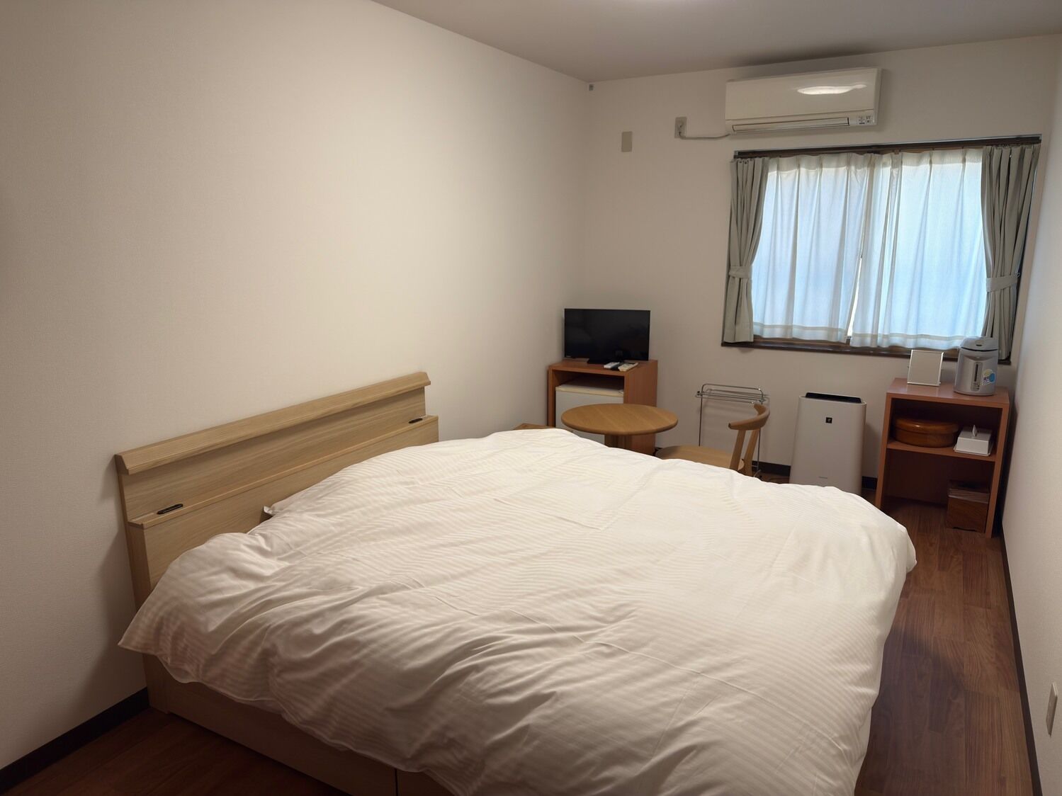 Habitación de hotel con cama, televisión, una mesita, una silla, radiador y ventana con cortinas.
