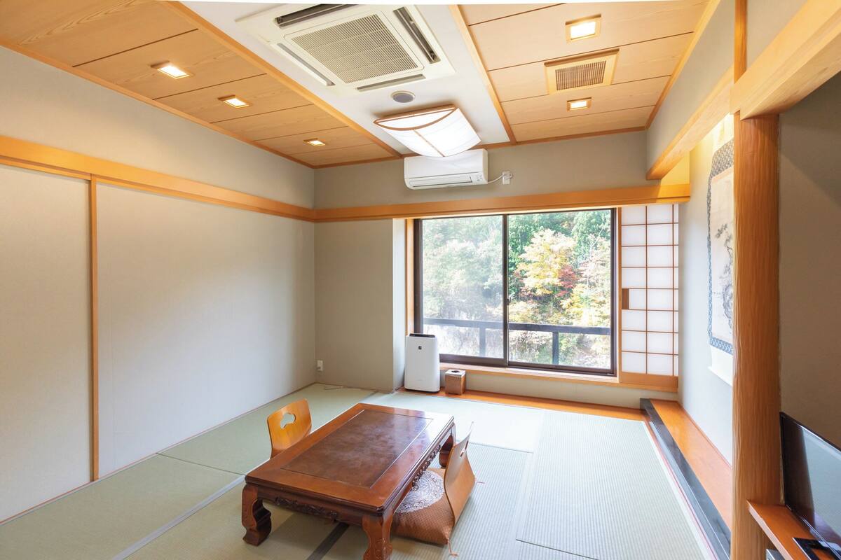 Una habitación con suelo de tatami, una mesa de madera, un televisor y un ventanal con vistas a árboles.