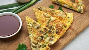A wooden cutting board with three pieces of fried food, likely a type of omelette or pancake, garnished with herbs. There is a small bowl of sauce to the left. - Indo Continental (Delhi)