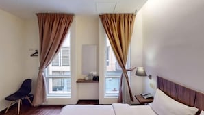 Una habitación de hotel con cama, una silla, una mesita de noche, lámpara y cortinas.
