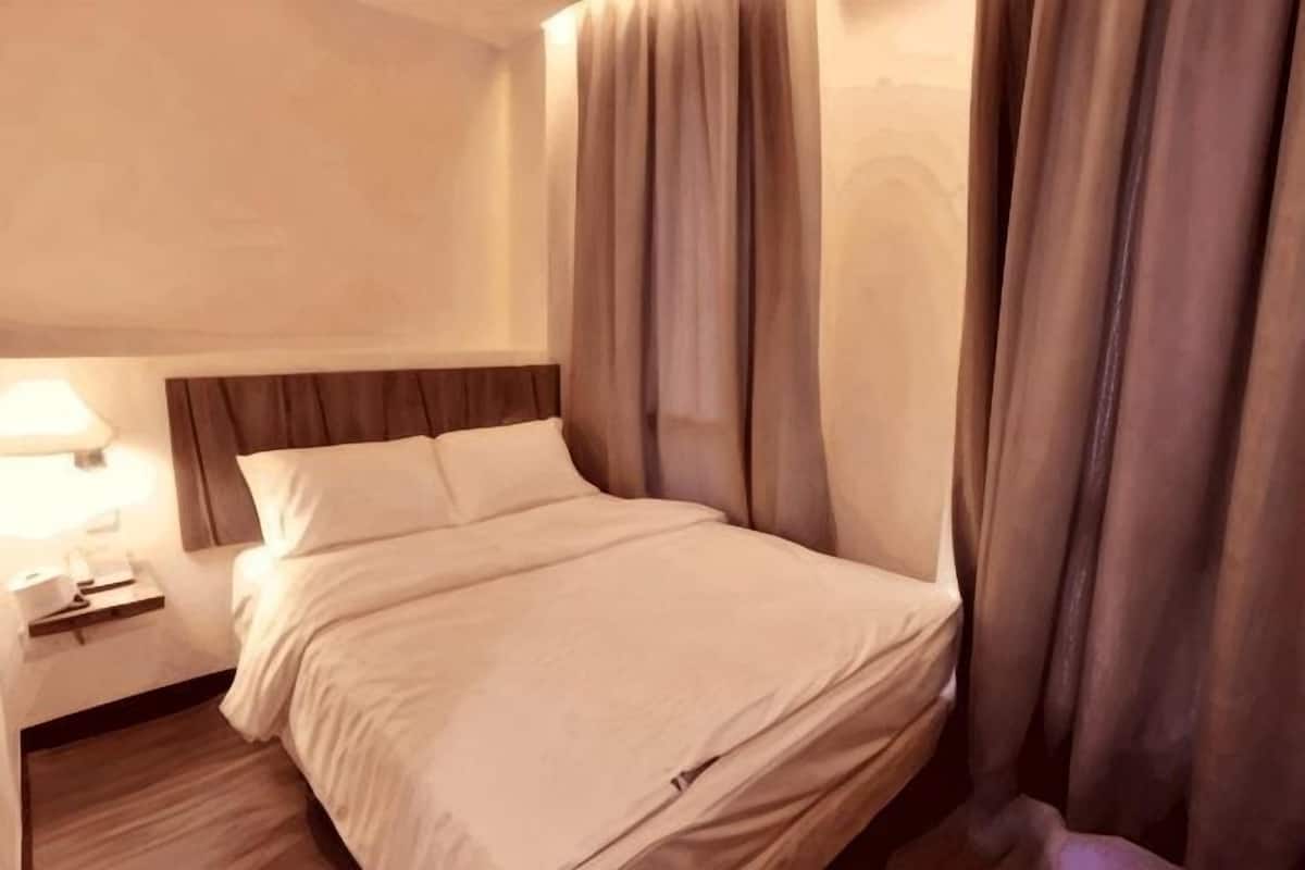 Una habitación de hotel con cama, cortinas, mesita de noche con lámpara y suelo de madera.
