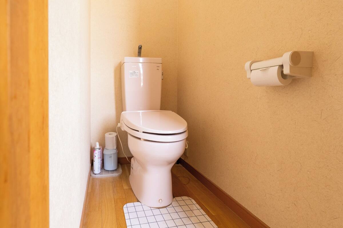 Une petite salle de bain avec des toilettes, un porte-rouleau de papier toilette et un produit de nettoyage posé sur le sol.