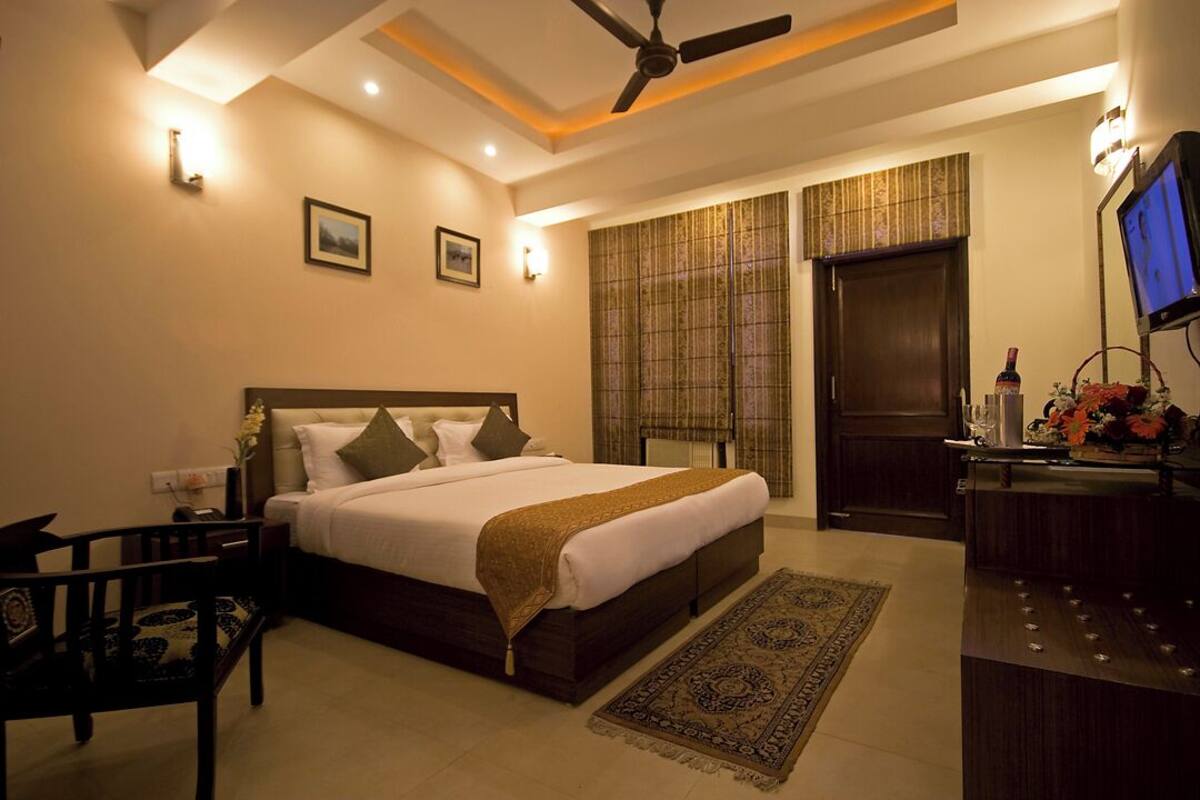 Quarto de hotel com cama, escrivaninha com televisão, ventilador de teto e janela com cortinas.