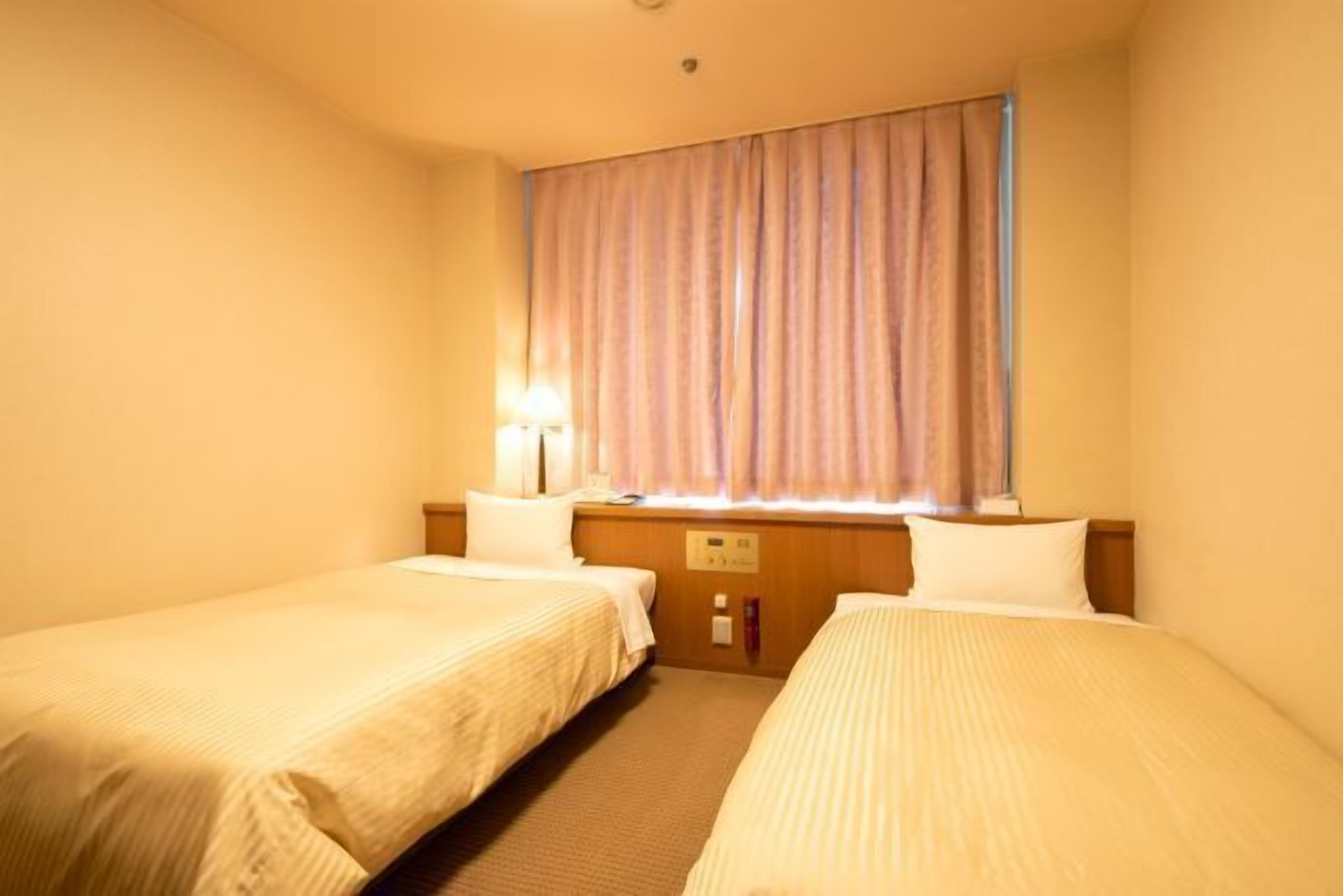 Ein Hotelzimmer mit zwei Einzelbetten, einem Holz-Kopfteil, einem Nachttisch mit Lampe und einem Fenster mit Vorhängen.