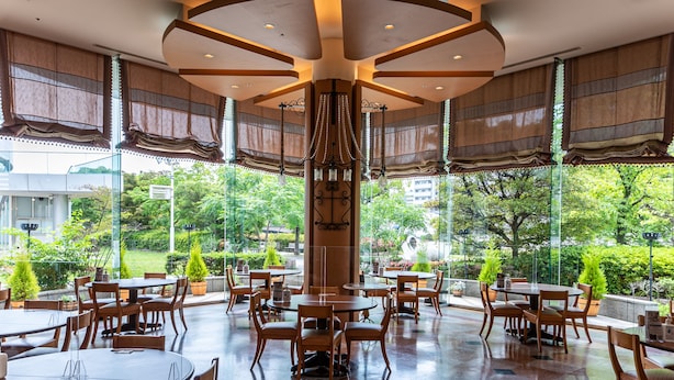 A spacious outdoor dining area with multiple tables and chairs, surrounded by large windows offering a view of greenery.