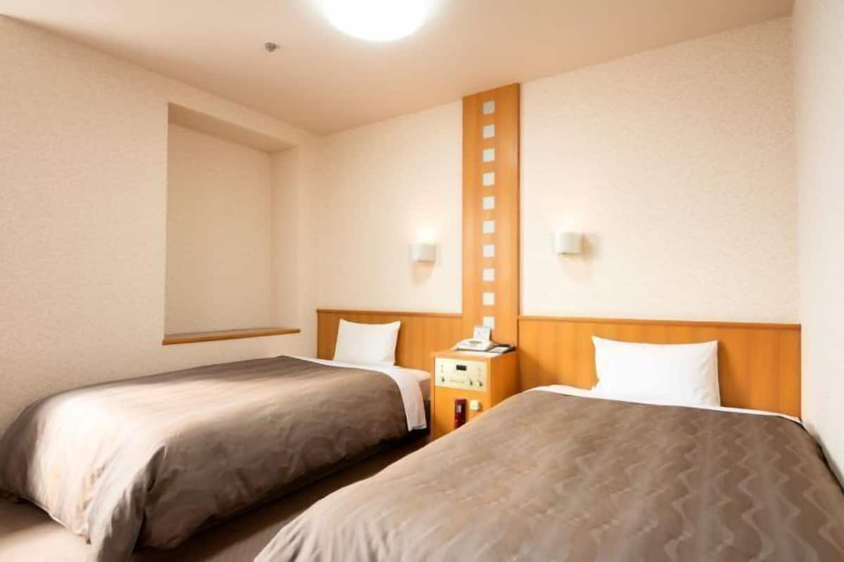 Hotellihuone, jossa on kaksi sänkyä, puinen pääty, yöpöytä lampun kanssa ja seinään kiinnitetty valaisin.