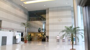 A spacious lobby with a high ceiling, large windows, and a modern design. There are potted palm trees and a reception desk.